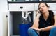 Woman sitting, calling for help as water leaks from pipe under sink into blue bucket.
