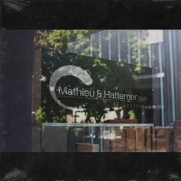 Firmenschild "Mathieu & Hattemer GbR IT Systems" an einer Glasfront mit Bäumen im Hintergrund.