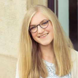 Eine lächelnde Frau mit Brille und langen blonden Haaren vor neutralem Hintergrund.