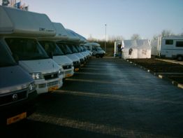 Reihe von Wohnmobilen auf einem Parkplatz, daneben steht ein weißes Zelt, Bäume im Hintergrund.