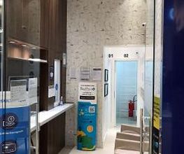 Corredor interior com cadeiras, placas informativas, paredes revestidas e porta de vidro.