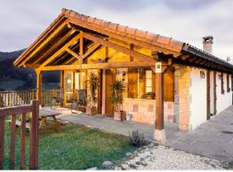 Casa rural de madera con techo a dos aguas, rodeada de campo y montañas al atardecer.