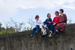 Four people sitting and smiling on a concrete wall against a blue sky background.