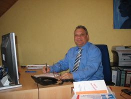 Mann im blauen Hemd sitzt an Schreibtisch mit Telefon und Computer, lächelt in die Kamera.