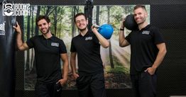 Drie mannen met sportkleding lachen in een fitnessruimte met een boksbal en gewichten.
