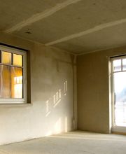 Leerer Raum mit unverputzten Wänden, einem kleinen Fenster und einer Glastür, Sonnenlicht fällt ein.