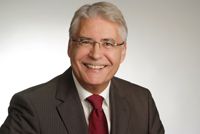 Ein lächelnder Mann mit Brille und Anzug, vor neutralem Hintergrund.