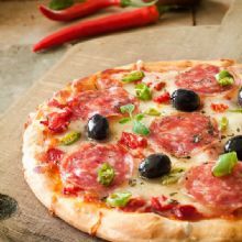 Pizza with salami, olives, and peppers on a wooden board with red chilies in the background.