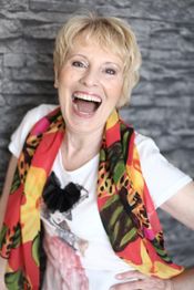 Ältere Frau lacht, trägt bunte Schaltuch und weißes T-Shirt, graue Steinwand im Hintergrund.