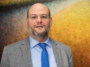 Ein Mann mit Brille und Bart in Anzug und blauer Krawatte vor einem gelben Hintergrund.