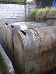 Drei rostige Metalltanks stehen nebeneinander in einem Hof mit Ziegelmauer und Pflanzen.