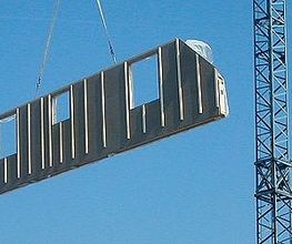 Kran hebt Wandsegment auf Baustelle in die Luft, blauer Himmel im Hintergrund.