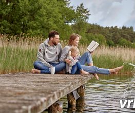 Familie sitzt auf dem Steg am See, liest und spielt im Wasser. Entspannter Sommertag in der Natur.
