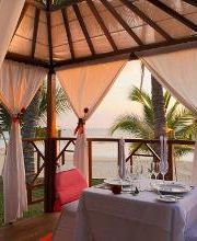 Mesa romántica bajo pérgola en la playa, al atardecer, con cortinas blancas y palmeras.