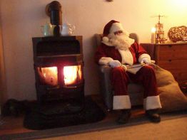 Weihnachtsmann sitzt neben einem brennenden Ofen in einem gemütlichen Raum, in einem Sessel.
