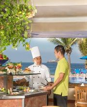 Chef y cliente conversando en un buffet al aire libre cerca del mar, con vegetación y palmeras.