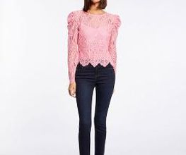 Mujer con blusa rosada de encaje y jeans oscuros, de pie en un fondo blanco.