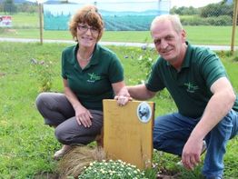 Ein Mann und eine Frau neben einem Holzkreuz auf einer Wiese, im Hintergrund ist ein Zaun.