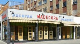 Fachada de tienda Madecora: puertas y parquet, con toldo amarillo y ventanas grandes.