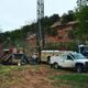 Drilling equipment and trucks set up near a rocky hillside and greenery.
