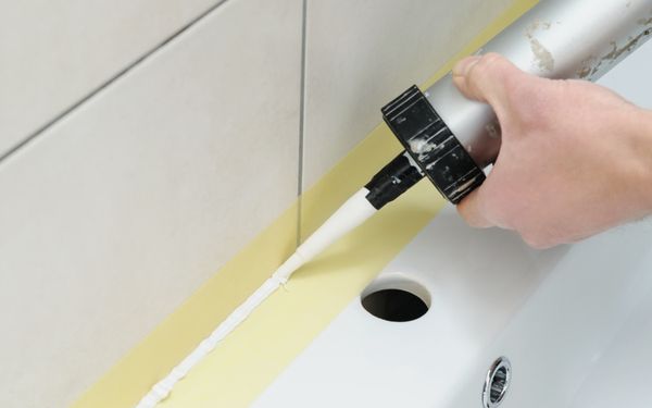 Am Übergang zwischen Waschbecken und Fliesenspiegel wird in dem mit Klebeband abgeklebten Bereich mithilfe einer Kartusche Silikon aufgetragen.