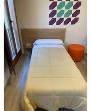 Cama individual con manta beige, pared decorada y taburete naranja en un dormitorio pequeño.