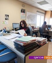 Oficina con dos personas trabajando en escritorios, rodeadas de pilas de documentos.