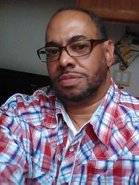 Man in glasses wearing a red and blue plaid shirt, looking at the camera indoors.