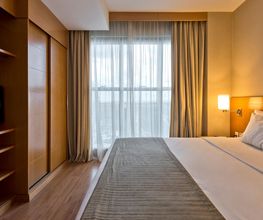 Quarto de hotel com cama de casal, TV, janela com cortinas e iluminação suave.