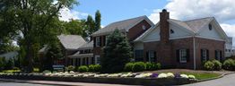 Brick house with a gabled roof, surrounded by trees and colorful flower beds under a blue sky.