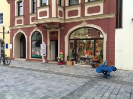 Straßenansicht eines Spielzeugladens mit Schaufensterdekoration, Fahrrad und blauem Spielgerät draußen.