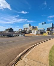 Fábrica industrial em dia ensolarado, céu azul com algumas nuvens, cercada por estrada asfaltada.