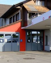 Tankstelle mit MIGROL-Logo, rote und weiße Gebäude, blauer Himmel im Hintergrund.