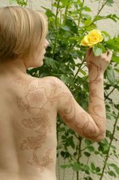 Person mit Blumen-Tattoo auf Rücken hält gelbe Rose vor blühender Pflanze.