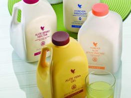 Vier Aloe-Getränkeflaschen auf einem Tisch mit einem Glas gefüllt mit einem gelblichen Getränk.
