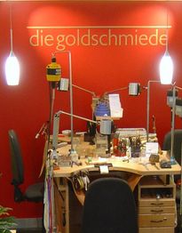 Goldschmiede-Werkbank vor roter Wand mit Schriftzug "die goldschmiede" und Hängelampen.