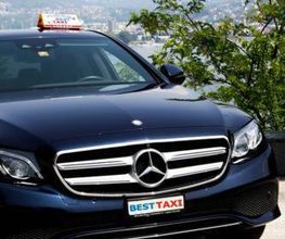 Schwarzes Taxi mit "Best Taxi"-Schild, parkend vor malerischem Hintergrund mit Bergen und Bäumen.