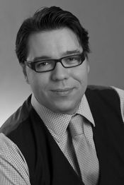 Eine lächelnde Person mit Brille, Hemd und Krawatte im Porträt, schwarz-weiß aufgenommen.