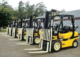 Cinco empilhadeiras amarelas estacionadas em linha em um pátio ao ar livre.