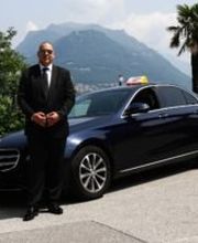 Ein Mann steht im Anzug neben einem schwarzen Mercedes-Benz vor Bergkulisse und Palmen.