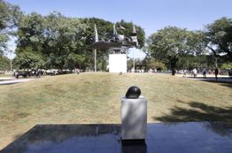 Escultura em parque com árvores, céu claro e pedestal em primeiro plano; pessoas passeiam ao fundo.