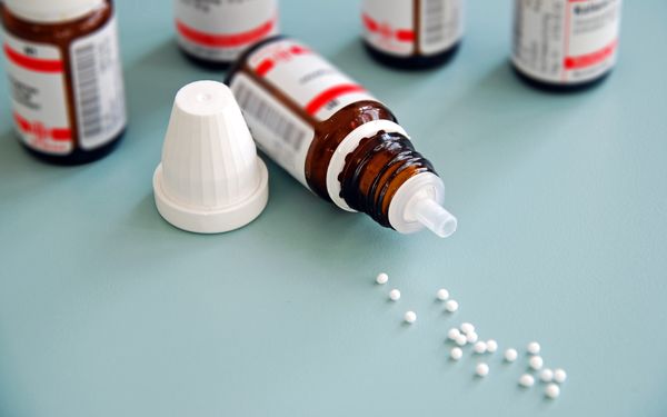 Ein umgefallenes Fläschchen Globuli liegt auf einem blauen Untergrund. Darum verstreut liegen kleine weiße Globuli Küglechen.