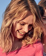 Zwei lächelnde Frauen in rosa Kleidung stehen eng zusammen vor blauem Himmel.