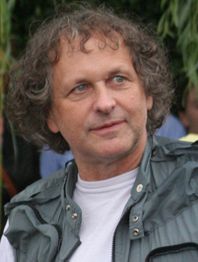 Ein Mann mit lockigem Haar im Freien trägt eine graue Jacke und ein weißes T-Shirt.