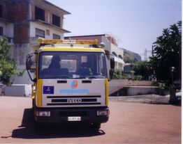 Camion giallo con logo Europ Assistance parcheggiato su strada, edificio sullo sfondo.