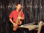 Mann spielt Saxophon und steht neben einem Keyboard vor einem glitzernden Hintergrund.