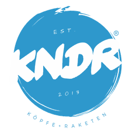Blaues Logo mit weißem "KNDR" Text, darunter "Köpfe + Raketen", etabliert 2013.