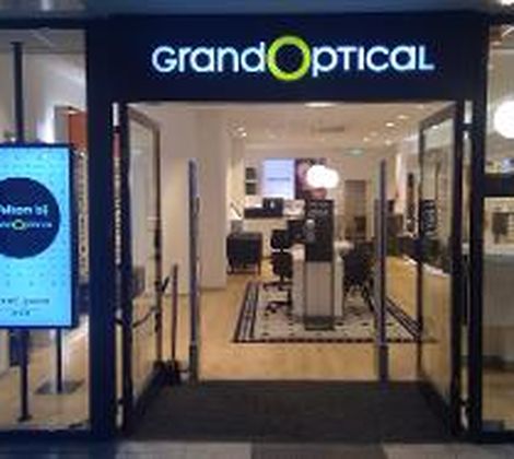 Winkelpui van een opticien met het bord "Grand Optical" en een welkomstdisplay aan de zijkant.