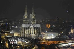Kölner Dom bei Nacht, von Lichtern der Stadt und beleuchteten Gebäuden umgeben.