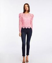 Mujer con blusa rosada de encaje y jeans oscuros, de pie en un fondo blanco.